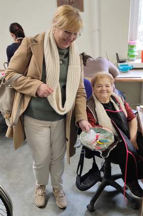 Pokaż zdjęcie: Dwie uśmiechnięte kobiety, uczestniczka WTZ siedzi przy stoliku i wykonuje ozdoby ceramiczne