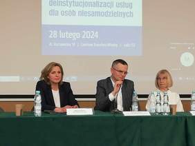 Pokaż zdjęcie: Sala konferencyjna, zdjęcie stołu prezydialnego przy którym siedzą trzy osoby, dwie kobiety i mężczyzna pośrodku, w tle slajd w prezentacji.