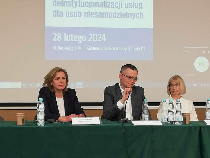 Pokaż zdjęcie: Sala konferencyjna, zdjęcie stołu prezydialnego przy którym siedzą trzy osoby, dwie kobiety i mężczyzna pośrodku, w tle slajd w prezentacji.