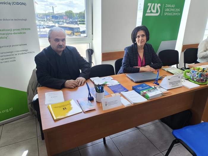 Pokaż zdjęcie: Przy stoliku na którym leżą ulotki i foldery siedzi mężczyzna i kobieta. Za nimi stoi baner z logo CIDON.