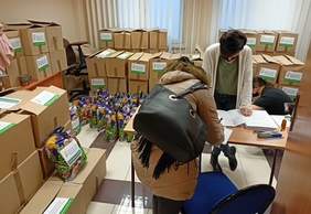 Pokaż zdjęcie: Pokój wypełniony paczkami i pudełkami z logotypem PFRON. Na pierwszym planie kobieta, która odbiera jedną z paczek