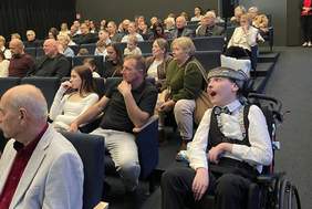 Pokaż zdjęcie: Grupa osób siedzących i stojących w sali podczas wydarzenia; na pierwszym planie osoby na wózkach inwalidzkich rozmawiają, w tle zgromadzona publiczność obserwuje spotkanie.