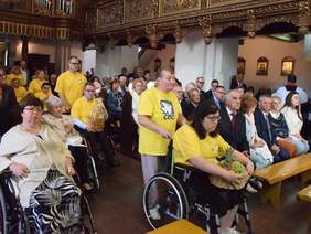 Pokaż zdjęcie: W kościele siedzi wielu wiernych. Wszyscy ubrani na galowo. Z przodu znajdują się osoby niepełnosprawne siedzące na wózkach inwalidzkich. U góry widać kawałki balkonu w złote zdobienia.