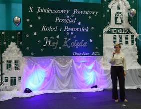 Pokaż zdjęcie: Kobieta stoi na scenie i przemawia do mikrofonu na tle świątecznej dekoracji z napisem „X Jubileuszowy Powiatowy Przegląd Kolęd i Pastorałek Hej, Kolęda, Długobórz 2026”. Scena ozdobiona jest jasnymi tkaninami, światełkami oraz elementami bożonarodzeniowej scenografii.
