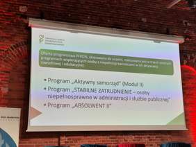 Pokaż zdjęcie: Na ścianie wisi duża tablica multimedialna. Wyświetlona jest na niej informacja dotycząca oferty programowej PFRON dla Uczelni oraz NGO. 
