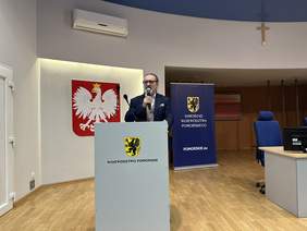 Pokaż zdjęcie: Z mównicą z godłem i napisem województwo pomorskie stoi mężczyzna w okularach, ubrany w granatową marynarkę i szarą kamizelkę. Trzyma mikrofon. Za nim duże godło polskie na białej podświetlanej ścianie.