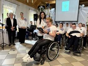 Pokaż zdjęcie: Grupa dorosłych osób z niepełnosprawnościami bierze udział w uroczystości; kobieta na wózku odczytuje tekst, obok niej stoi asystentka, za nimi siedzi kilkanaście osób w białych koszulach, w tle ekran z prezentacją i dekoracje.