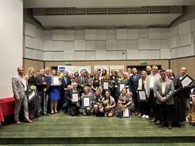 Pokaż zdjęcie: Na scenie stoi wiele elegancko ubranych osób, trzymają dyplomy, kwiaty i statuetki. Wszyscy są szczęśliwi.