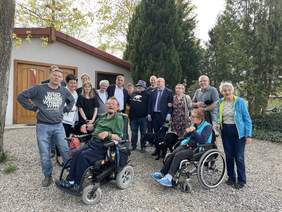 Pokaż zdjęcie: Przed niską kapliczką stoi grupa uśmiechających się osób. Są tu osoby na wózkach inwalidzkich. Po środku stoi ksiądz w czarnej koszulce z napisem PAPA BEAR. Obok niego dwaj mężczyźni ubrani na galowo i pies asystujący,