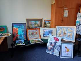 Pokaż zdjęcie: W pokoju z niebieską wykładziną wyeksponowane są prace plastyczne. Kategoria malarstwo.