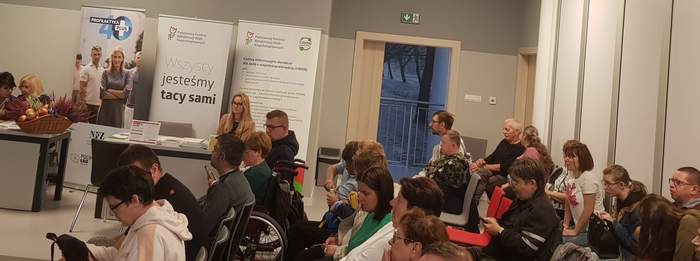 Pokaż zdjęcie: Duża grupa osób siedzi w sali, jedna z nich porusza się na wózku. Za stolikiem na tle baneru PFRON oraz CIDON siedzi kobieta reprezentująca Oddział Wielkopolski PFRON. Na stoliku ułożone są materiały informacyjne promujące programy i zadania PFRON.