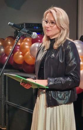 Pokaż zdjęcie: Kobieta w okularach z zieloną teczką w dłoniach przemawia do mikrofonu. W tle różnokolorowe balony.