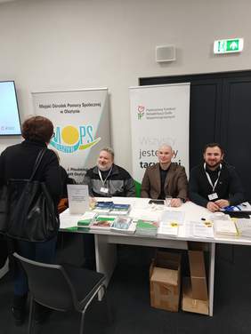 Pokaż zdjęcie: Trzech mężczyzn siedzi przy stole z licznymi ulotkami, informatorami oraz broszurami. Przed nimi kobieta stojąca tyłem do aparatu.