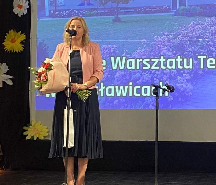 Pokaż zdjęcie: Na scenie przed mikrofonem stoi kobieta ubrana w granatową sukienkę i łososiową marynarkę. W ręku trzyma bukiet kwiatów.  