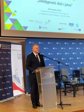 Pokaż zdjęcie: Przy mównicy stoi siwy mężczyzna w ciemnym garniturze.