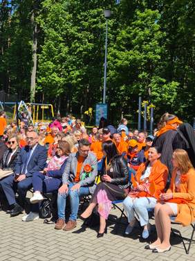 Pokaż zdjęcie: Zdjęcie zaproszonych gości siedzących na krzesełkach, wiele osób ubranych jest na pomarańczowo.
