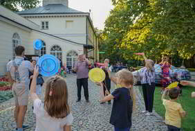 Kilka osób: dzieci i dorośli bawią się kręcąc kolorowe talerze na patykach. Stoją na chodniku wyłożonym kostką brukową. Z lewej strony widzimy zarys budynków a z prawej zielone, liściaste drzewa.