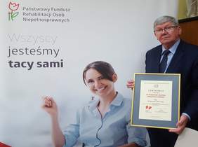 Pokaż zdjęcie: na zdjęciu:  jedno z pomieszczeń Oddziału Łódzkiego PFRON w tle baner z logo PFRON i napisem wszyscy jesteśmy tacy sami. Z prawej strony Dyrektor Oddziału Łódzkiego PFRON ubrany w garnitur trzyma  oprawiony w ramce Certyfikat Afirmatora Ruchu Inn