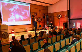Pokaż zdjęcie: Sala konferencyjna, scena z prelegentem, krzesełka w układzie kinowym, widać osoby siedzące na widowni. 