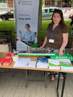 Pokaż zdjęcie: Kobieta w okularach słonecznych na głowie stoi za stołem na stoisku promocyjnym oddziału lubelskiego PFRON, na stole leżą materiały promocyjne