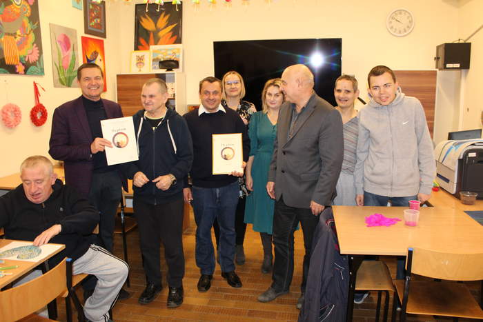 Pokaż zdjęcie: Zdjęcie sali  w warsztacie terapii zajęciowej, na tle tablicy stoi mieszana grupa osób. Dwie osoby trzymają w ręku dyplomy