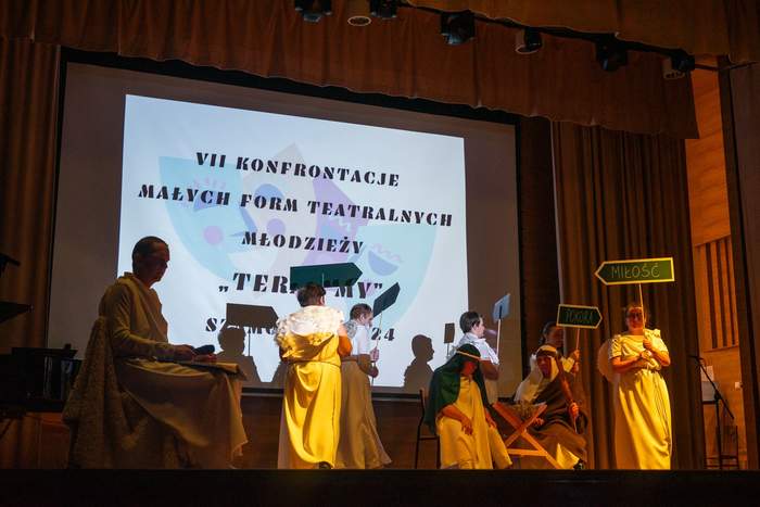 Pokaż zdjęcie: Na scenie teatralnej znajduje się grupa sześciu osób przebranych za anioły. Dwie dziewczyny trzymają w dłoniach tabliczki będące drogowskazami z napisem: miłość oraz pokora. Na scenie widoczny jest również żłóbek, przy którym siedzą osoby przebrane za Jezusa oraz Maryję. W tle, na dużym ekranie wyświetlona jest nazwa wydarzenia, czyli: VII Konfrontacje Małych Form Teatralnych Młodzieży „Teraz My” w Szamotułach.
