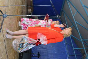 Pokaż zdjęcie: Na scenie stoją dwie kobiety, jedna z nich ubrana w czerwona sukienkę trzyma kosz z cukierkami w ręku i przemawia