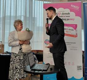 Pokaż zdjęcie: Mężczyzna w garniturze przemawia do mikrofonu, patrząc na stojącą obok niego kobietę. Kobieta w jasnej marynarce i spódnicy o wzorze w czarne i białe koła uśmiecha się i trzyma bukiet kwiatów. Obok nich znajduje się stolik z otwartym laptopem. W tle widoczny jest baner z informacjami o suplemencie diety CDS22-formula.