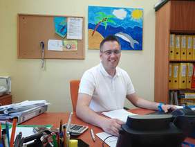 Pokaż zdjęcie: Pan siedzi w pracy za biurkiem przed komputerem. Uśmiecha się do aparatu fotograficznego. Na biurku leżą różne dokumenty i sprzęty biurowe. Na ścianach w tle wiszą kolorowe obrazki. 