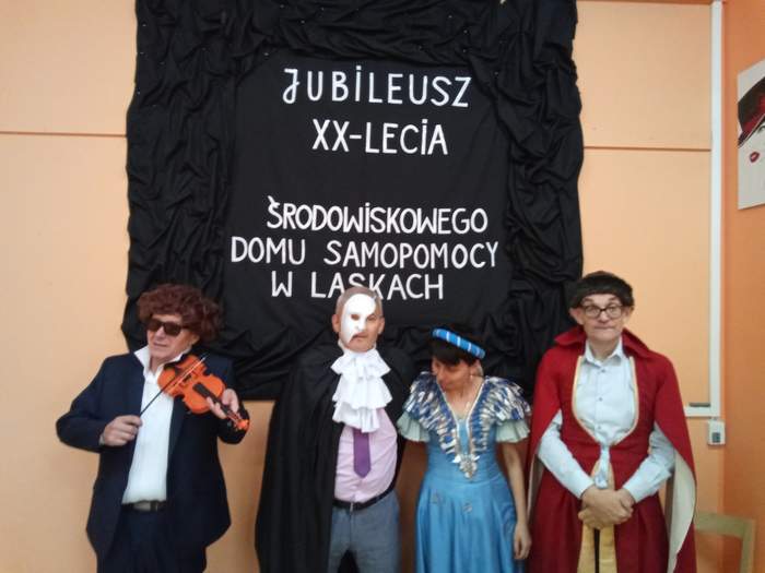 Pokaż zdjęcie: Na tle czarnej kurtyny z napisem jubileusz 20 lecia, stoją 4 osoby przebrane w starodawne stroje