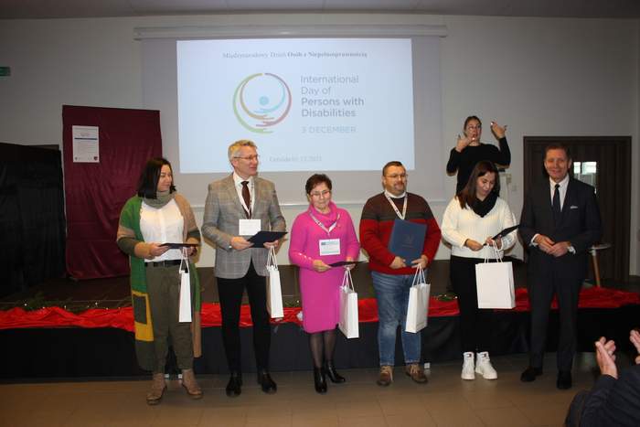 Pokaż zdjęcie: Kilka osób stoi na środku sali i trzyma dyplomy, za nimi stoi tłumacz języka migowego