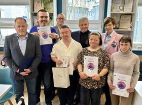 Zdjęcie grupowe ośmiu osób, z których część trzyma pamiątkowe dyplomy laureatów i uczestników XXII edycji wojewódzkiego etapu Konkursu Plastycznego „Sztuka Osób Niepełnosprawnych”.