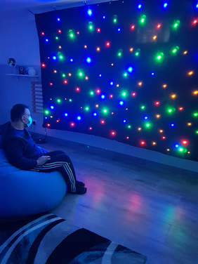 Pokaż zdjęcie: W ciemnym pokoju na miękkim siedzisku siedzi mężczyzna w maseczke, patrzy się na granatową ścianę na której wyświetlają się kolorowe kropki. Kropki sa podświetlone, różnej wielkości w kolorze zielonym, niebieskim i czerwonym.