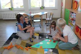 Pokaż zdjęcie: W sali na kolanach u pani siedzi chłopiec, obok druga dziewczynka z Panią. Przed nimi na dywanie leży zielony latawiec,który ozdabiaja kolorywymi kwiatkami