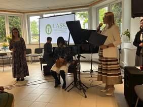 Pokaż zdjęcie: W centralnym punkcie zdjęcia siedzi niewidoma podopieczna OSW grająca na ukulele. Za nią, przy fortepianie siedzi kobieta. Po lewej stronie stoi podopieczna z mikrofonem w ręce. Po prawej stronie stoi pani dyrektor OSW trzymająca w rękach mikrofon i podziękowania.