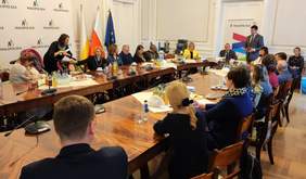 Pokaż zdjęcie: Uczestnicy spotkania siedzą przy dużym owalnym stole. Wszyscy słuchają mężczyzny, który stoi na środku i przemawia.