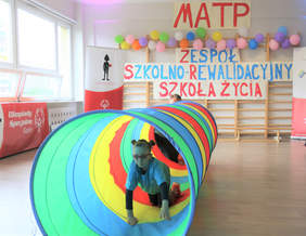 Pokaż zdjęcie: Na sali gimnastycznej jest rozłożony długi, kolorowy tunel przez który przechodzi dziewczynka w okularach