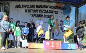 Pokaż zdjęcie: Grupa osób stoi na scenie pod zadaszeniem podczas ceremonii wręczenia nagród. W tle znajduje się baner z napisem: „Ogólnopolskie Wiosenne Mistrzostwa w Biegach Przełajowych ‘Sprawni Razem’”. Na podium stoją trzy młode zawodniczki z medalami i nagrodami – na najwyższym stopniu dziewczyna w zielonej koszulce, na drugim dziewczyna w niebieskiej, a na trzecim zawodniczka w białej koszulce. Obok nich znajdują się organizatorzy i inni uczestnicy, którzy gratulują i obserwują wydarzenie. Scena udekorowana jest również logo miasta Nowogard i sponsorów.