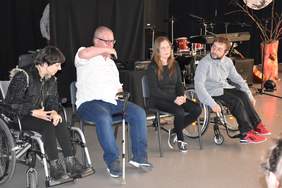 Pokaż zdjęcie: Na scenie w półokręgu na krzesłach lub wózkach siedzą osoby, zdjęcie przedstawia 4 osoby, jedna z nich mówi do mikrofonu.