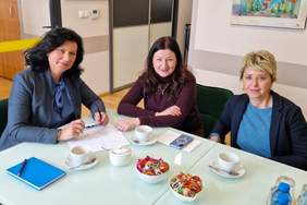 Pokaż zdjęcie: Przy stole siedzą trzy elegancko ubrane kobiety. Na stole są rozłożone dokumenty projektowe.