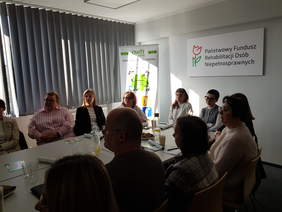 Pokaż zdjęcie: Zdjęcie przedstawiające spotkanie w sali konferencyjnej. W pomieszczeniu widać około kilkanaście osób (kobiety i mężczyźni). Ubrani są w stroje codzienne i półformalne (swetry, koszule, marynarki). Wszyscy siedzą i są zwróceni w stronę osoby prowadzącej spotkanie. Na stole leżą notatniki i ulotki; stoją też szklanki, butelka wody i dzbanek z napojem. Na ścianie po prawej stronie jest tablica z napisem: „Państwowy Fundusz Rehabilitacji Osób Niepełnosprawnych” oraz logo z czerwonym tulipanem. Po lewej stronie stoi roll-up z napisem „OWIT” i grafiką w zielonych barwach. Po lewej stronie sali są pionowe żaluzje koloru szarego zasłaniające okno. 
