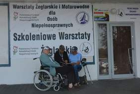 Pokaż zdjęcie: 3 panów pod banerem informacyjnym o Warsztatach żeglarskich dla osób niepełnosprawnych. Pan z lewej strony siedzi na wózku inwalidzkim, przy mężczyźnie z prawej strony stoją kule. Mężczyźni siedzący w środku i po prawej nachylają się nad l
