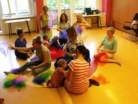 Pokaż zdjęcie: Grupa dzieci i dorosłych siedząca na podłodze podrzucająca chustki tiulowe