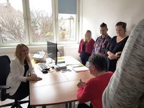 Pokaż zdjęcie: Przy biurku siedzi kobieta w białej marynarce, a naprzeciwko niej osoba w czerwonym swetrze, widoczna od tyłu. Za biurkiem stoją trzy dodatkowe osoby, które obserwują rozmowę. Scena przedstawia instruktaż dotyczący udzielania informacji w biurze.