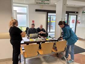 Pokaż zdjęcie: Dwóch pracowników PFRON siedzi przy stanowisku informacyjnym. Na stole znajdują się laptop oraz materiały informacyjne PFRON. Pracownicy rozmawiają z dwiema kobietami.