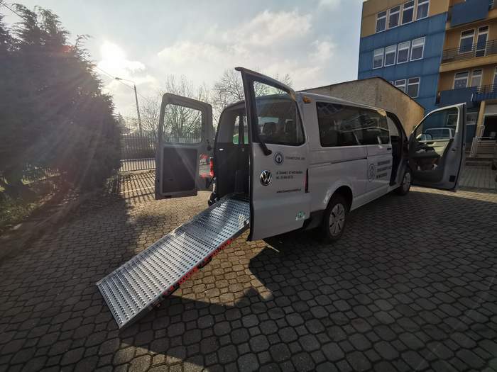 Pokaż zdjęcie: Na wypuzlowanym parkingu stoi biały bus ma otwarte drzwi po bokach z tyłu pojazdu jest wysunięta srebrna platforma