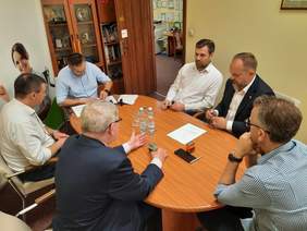 Pokaż zdjęcie: Przy owalnym stole siedzi sześciu mężczyzn, którzy ze soba rozmawiają. Jeden z panów jest pochylony nad dokumentami, do których przystawia pieczątkę. 