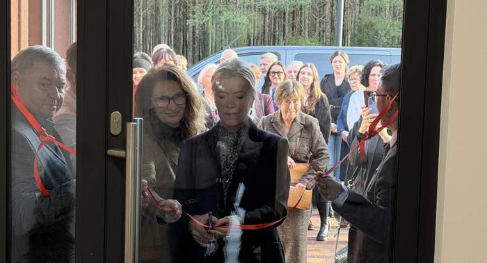 Pokaż zdjęcie: Uroczyste przecięcie czerwonej wstęgi przy wejściu do budynku. Kilka osób stoi przy drzwiach, jedna osoba trzyma nożyczki. W tle zgromadzeni uczestnicy wydarzenia, las i zaparkowany samochód.