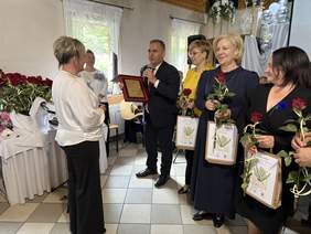 Pokaż zdjęcie: Uroczystość jubileuszowa – mężczyzna wręcza kobiecie tablicę pamiątkową, obok kobiety z różami i torbami z napisem „Jubileusz 50-lecia 1973–2023”, w tle dekoracje i mężczyzna z gitarą.