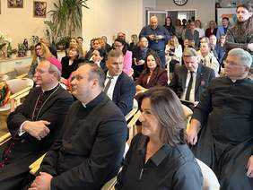Pokaż zdjęcie: Uczestnicy uroczystości siedzą w sali, zwróceni w stronę sceny. W pierwszych rzędach widoczni są duchowni, w tym biskup, oraz pozostali goście. Część osób stoi z tyłu sali.